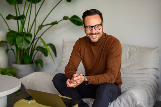 istock Happy businessman with glasses looking away thoughtfully while working over laptop on sofa in office 2157651041