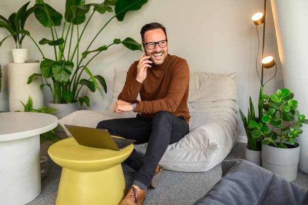 istock Happy male computer engineer discussing over phone call while working over laptop on sofa in office 2157651077