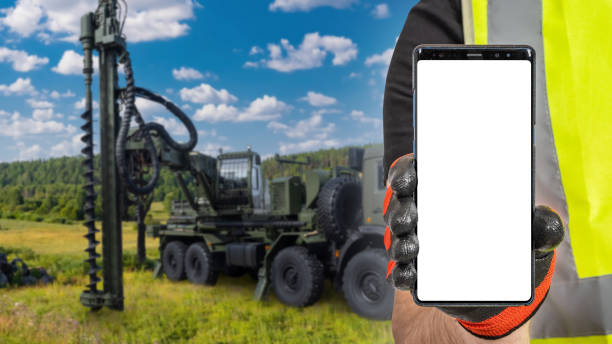 istock Construction business. Phone in builder hand. Machine for drilling wells in ground. Phone with blank screen. Drilling well for water production. Development water wells during construction. Art focus 2157671495