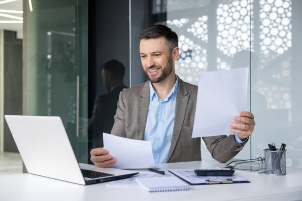 istock Smiling businessman in office holding documents and working on laptop 2157673677