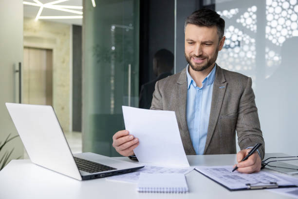 istock Smiling businessman working at desk with laptop, reviewing documents and taking notes 2157675267