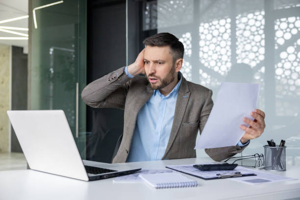 istock Stressed businessman reviewing documents in modern office while working on laptop 2157675472