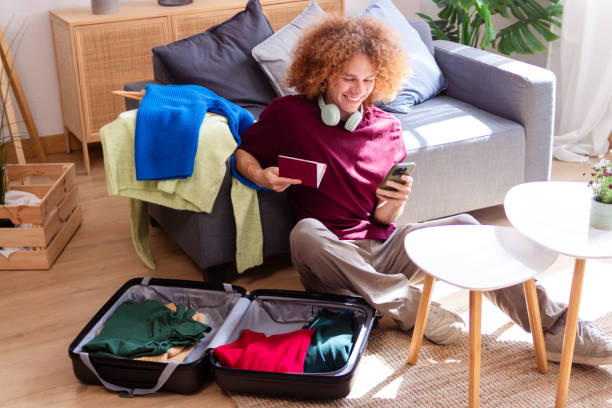 istock Young man holding passport and tickets using smartphone while packing suitcase at home 2157680857