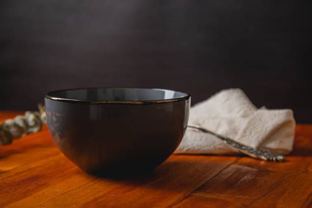 a dark scene consisting of a dark empty gray ceramic bowl, that can be used as mockup, over a wooden talbe with a colth and eucalyptus branch behind it