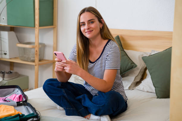 istock Smiling young woman using smartphone while sitting on bed with suitcase open 2157713254