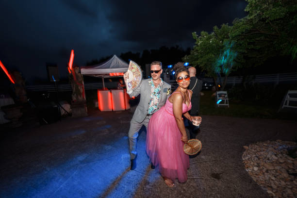 A multiracial group of adults have fun dancing to music performed by a DJ while attending an outdoor wedding reception at night.