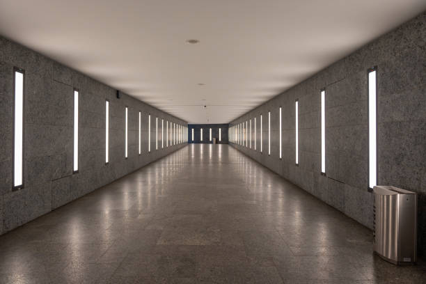 A long, empty hallway with a silver trash can in the middle. The walls are made of stone and the floor is tiled. The lighting is bright and the hallway appears to be empty