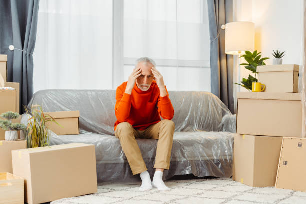 Tired senior man sitting on sofa feeling sad preparing unpacking cardboard boxes