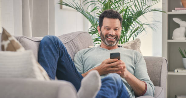 istock Internet meme, smartphone and relax with man on sofa for social media, laughing and text message. Happy, smile and technology with guy in living room at home for funny joke, connection or digital app 2157720595