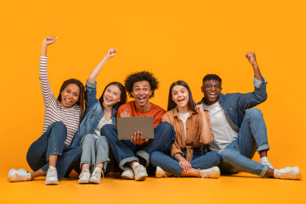 istock Seated friends celebrating with a laptop on orange background 2157722446