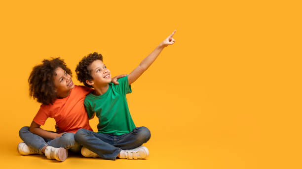 Two Children Sitting on Yellow Background Pointing