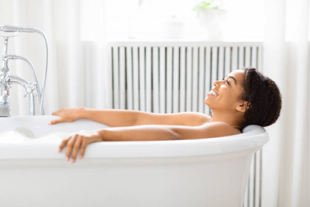 Woman enjoys serene moment in tub at home Serene young African American woman relaxing in a filled bathtub, looking up with closed eyes in a pristine bathroom drenched stock pictures, royalty-free photos & images