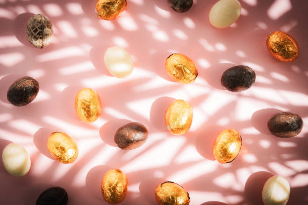 Beautiful atmospheric view of a mixture of chocolate eggs lying scattered on a pink background with soft shadows from a palm branch, flat lay close-up. World Chocolate Day concept.