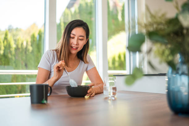 Successful Asian business woman sitting at a dining table and eating breakfast alone, thinking of something pleasant and smiling before her day starts. A glass of water and a cup of tea next to the bowl with cereal she is eating from.