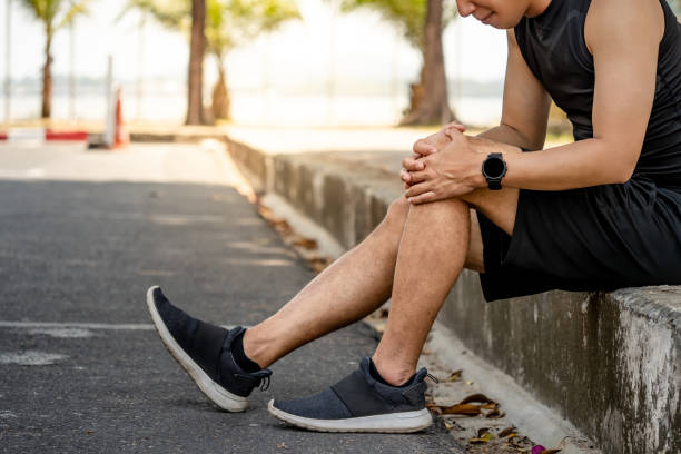 Athlete man feeling knee pain or rheumatoid arthritis while sitting on footpath in the park. Asian male runner suffering from acute knee joint inflammation symptom