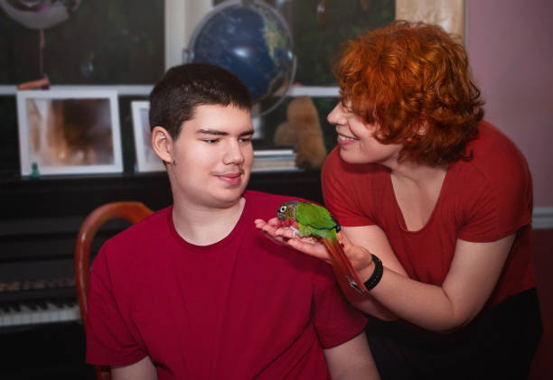 Mother is presenting a green-cheeked parakeet perched on her hand to her 18-year-old son indoors. The warm scene highlights the importance of the interaction between the teenage boy and his mother. The indoor setting enhances the engaging atmosphere as they share this moment with the pet bird.