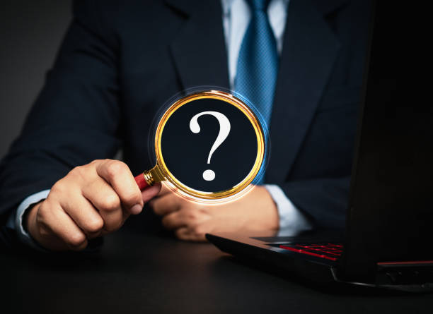 istock Businessman holding a magnifying glass with a white question mark symbol while sitting at a desk. 2157783785