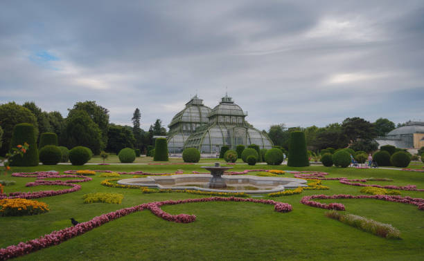 Vienna, Austria - 27 July 2023: beautiful old greenhouse orangery in the park of Schönbrunn palace, founded in 1882. The view on park with buildings, trees and colorful flowers evening sunset time.