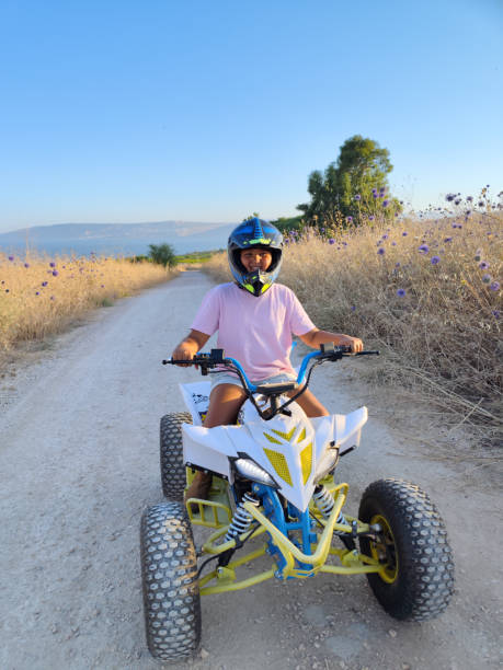 In this image, a young child is riding an ATV on a dirt path surrounded by dry, grassy terrain and vibrant purple wildflowers. The child is wearing a protective helmet and a light pink T-shirt, exuding a sense of joy and excitement as they navigate the trail. The ATV, built for off-road adventures, is prominently displayed with its large wheels and robust frame. The background features a clear blue sky and distant hills, adding to the sense of freedom and exploration. The scene captures a moment of youthful adventure and the thrill of outdoor activities in a picturesque, natural setting.