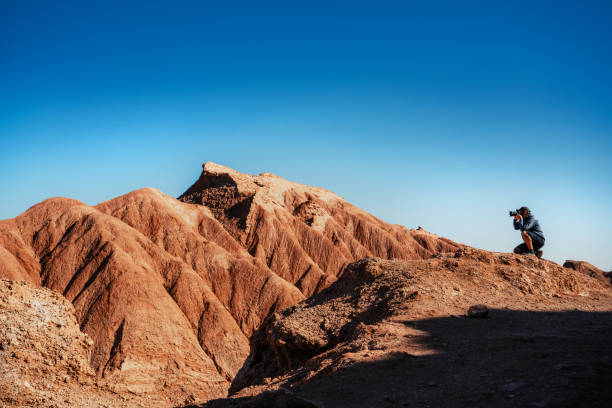 A man in casual attire takes photographs of unique rock sculptures in Vallecito, Atacama Desert, Chile, showcasing the stunning natural beauty of the dry landscape.