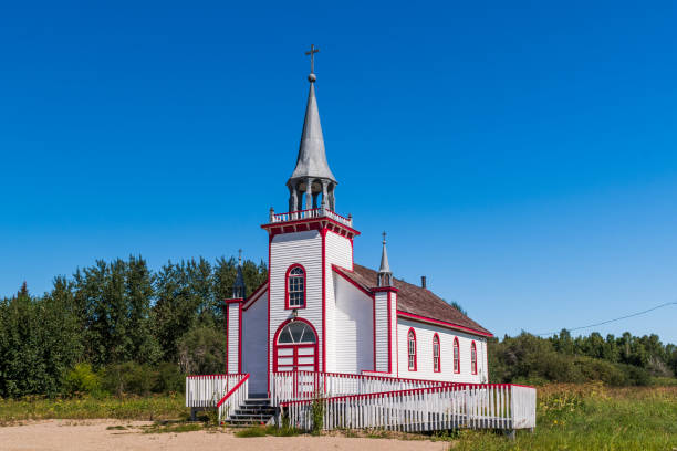 Small white church in Davisburg, featuring a dark roof, red door, and arched windows. The building is set in an open field with dry grass, flanked by two tall evergreen trees under a clear blue sky.
