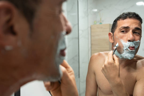 istock Young Adult Man Focused on Shaving Routine in Bathroom Mirror With focused expression and lips pressed. Shirtless man carefully shaves cheek in bathroom, prioritizing grooming. 2157854896