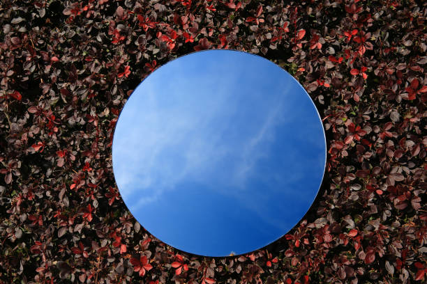 istock Round mirror on red and brown leaves reflecting sky, top view 2157875915