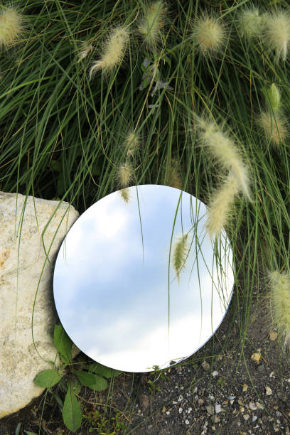 istock Round mirror on ground near grass and spikelets reflecting light blue sky 2157876000