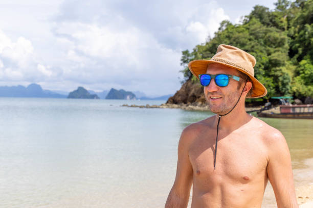 A man in a hat and sunglasses smiles while standing on a tropical beach, with calm waters and scenic islands in the background.