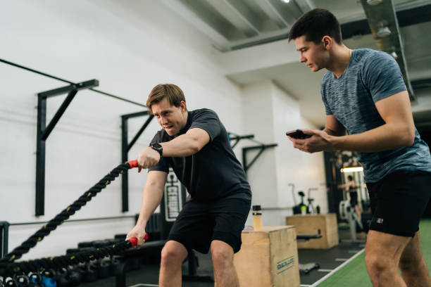 istock Low-angle view of personal trainer coaching beginner sportsman to perform battle ropes exercise in modern fitness gym. Concept of workout training coach business 2157917816