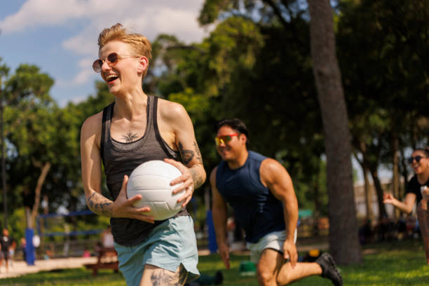 Non-binary White person holding volleyball ball, vigorously sprinting across sunlit grassy park, followed by Hispanic Latino man. Scene filled with laughter, motion, and teamwork, captured on a sunny day, showcasing a moment of active recreation among diverse friends.