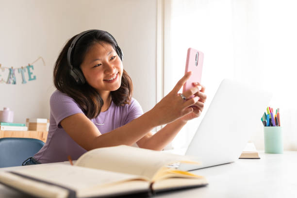 istock Young female Asian college student distracted by social media while studying at home. Taking selfie, video call. 2157938485