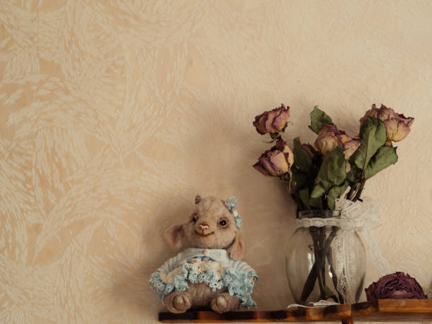 An adorable Teddy Lamb is wearing a blue lace-trimmed dress, accompanied by a bouquet of dried roses, both resting on a rustic wooden shelf. The Teddy Lamb's intricate dress and the delicate petals of the dried roses evoke a sense of nostalgia and timeless beauty. The rustic wooden shelf adds a touch of warmth and character to the composition, enhancing the vintage aesthetic in a nursery bedroom. The soft, muted tones of the scene create a serene and elegant atmosphere. This charming display blends the old-world charm of vintage decor with the refined artistry of modern creators.