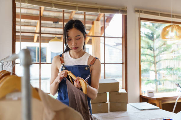 Young asian online merchant women using hanger to hanging new collection sweater on clothes rack while working about business shopping online to selling and shipping delivery product at home office. jóvenes mujeres asiáticas comerciantes en línea que usan la percha para colgar el suéter de la nueva colección en el perchero mientras trabajan en las compras comerciales en línea para vender y enviar el producto de entrega en la oficina en casa