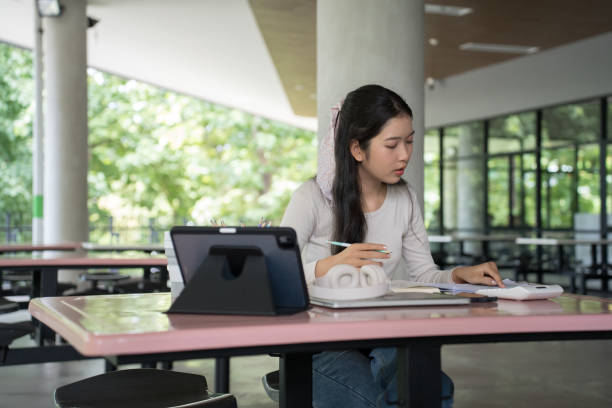 istock Young asian student woman checking business education paperwork and taking notes in notebook while watching video to studying and learning about business knowledge in online class at university 2157952200