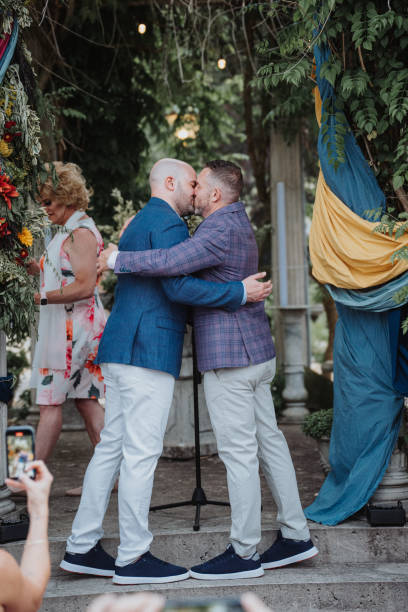 Gay couple kiss at wedding ceremony A gay couple leans in to exchange a kiss after saying their vows at their outdoor wedding ceremony attended by supportive family and friends. gay wedding kiss stock pictures, royalty-free photos & images