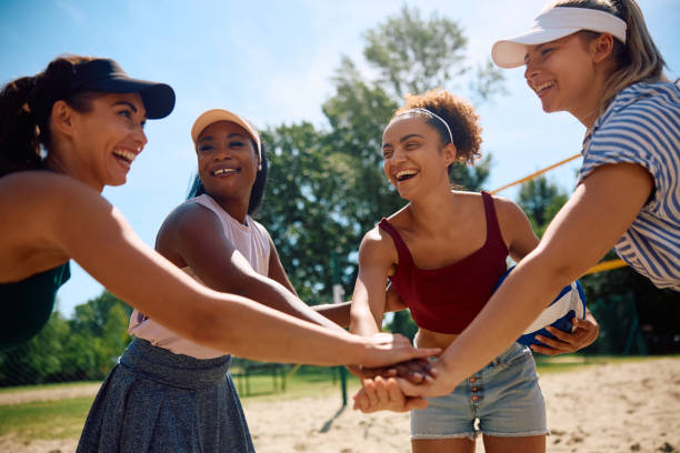 Cheerful female friends gathering hands in unity while playing beach volleyball.
