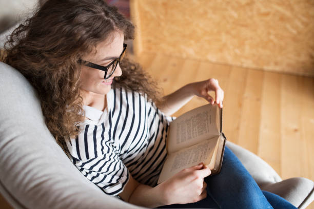 istock Female student sitting in armchair, reading book, studying for final exam at university. Beautiful woman relaxing at home. 2157969434