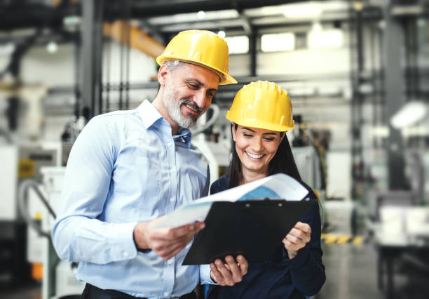 istock Two project managers standing in modern industrial factory, looking document in clipboard. Manufacturing facility with robotics, robotic arms and automation. Storing products and materials in warehouse. 2157971785
