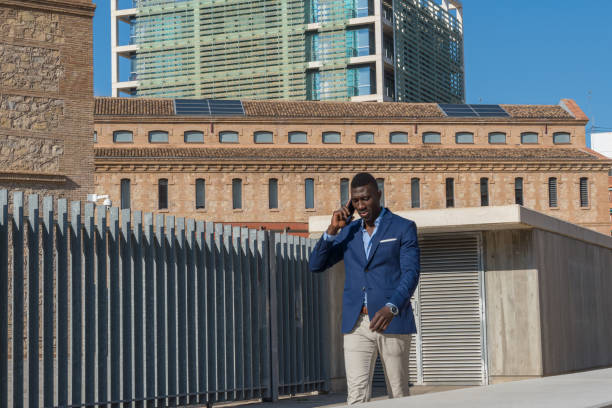Stylish African American Businessman Talking on Smartphone While Walking Between Modern Office Buildings in Urban Financial District.
