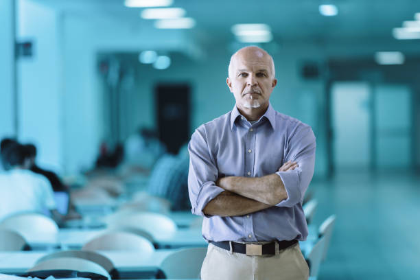 Portrait of a confident professor posing with crossed arms in a university hallway, students are sitting in the background, blurred, studying