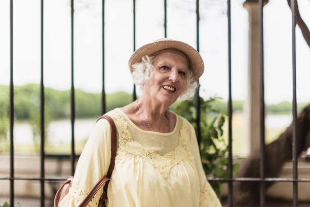 An elderly woman wearing a hat and a light yellow shirt is smiling while standing outdoors. The background shows greenery and a black fence, suggesting a park or garden setting.