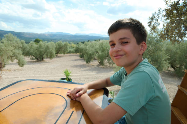 A young boy with a happy expression sitting at a table outdoors, surrounded by olive trees with mountains in the background. Sunny day with clear skies.