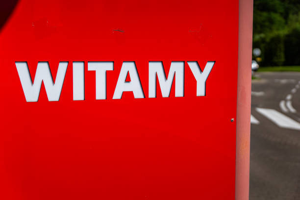 A close-up shot of a red road sign with the word Witamy written in white letters. The sign is mounted on a metal pole and is visible against a blurry background of a paved road and green grass.