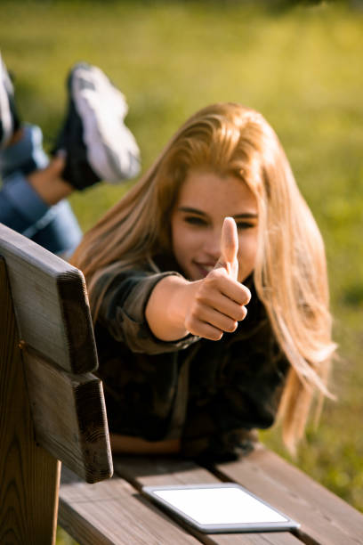 Young blonde woman lies on a park bench, giving a cheerful thumbs up with a tablet by her side, exuding a positive and relaxed vibe