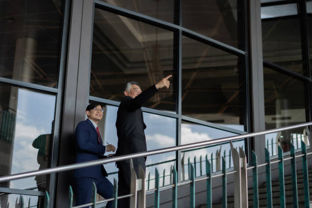 istock Two men in suits are standing on a railing, one pointing to something. Scene is professional and serious 2158039427