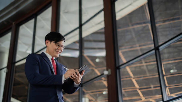 istock A man in a suit is looking at a tablet while standing in a large room. He is focused on the tablet, possibly checking emails or working on a project. The room is well-lit and has large windows 2158043583