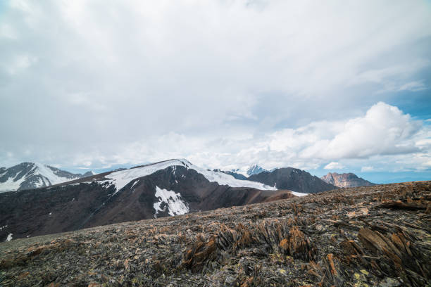 istock Sharp stones on high stony mountain pass with panoramic view to big glacier tongue among rockies and large snow-capped pinnacle in cloudy sky. Spectacular alpine landscape in high rocky snow mountains 2158044757