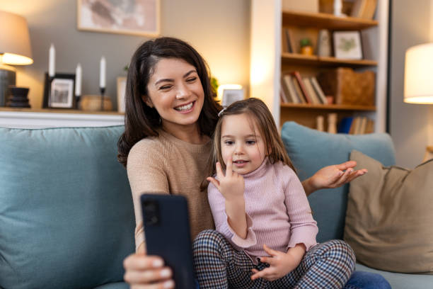 istock Smiling mother and daughter on video call using digital phone at home. while making video call and waving hello sitting on sofa. Woman and her cute little kid doing video conference with grandparents. 2158066155