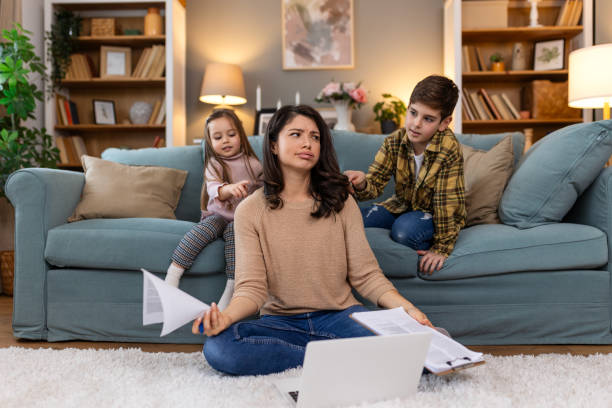 istock Tired young mother sitting on sofa and working with laptop and documents while little kids, her daughter and son are having fun on the sofa and making noise 2158066166